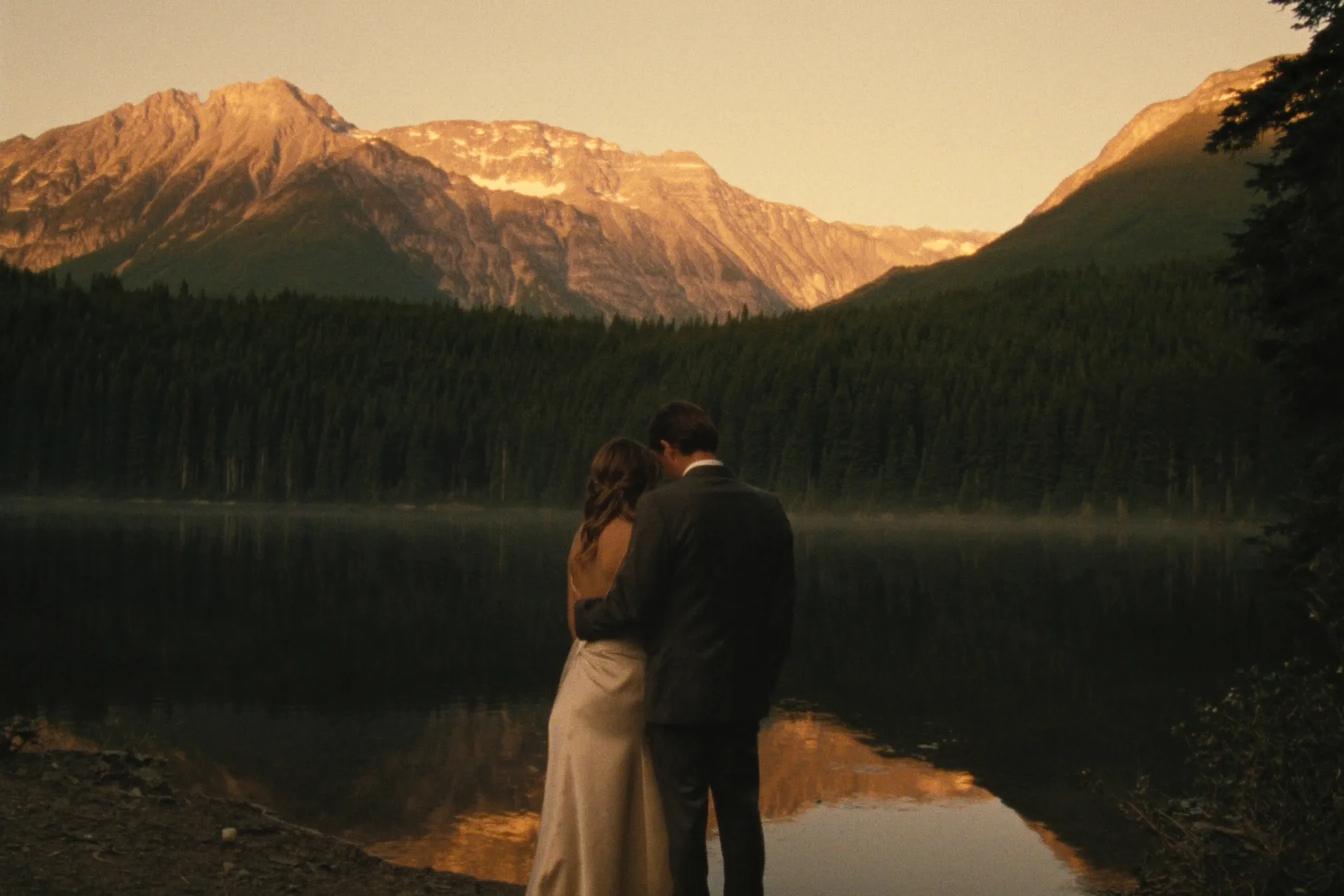 A couple at the edge of a still alpine lake at golden hour, peaks going rose