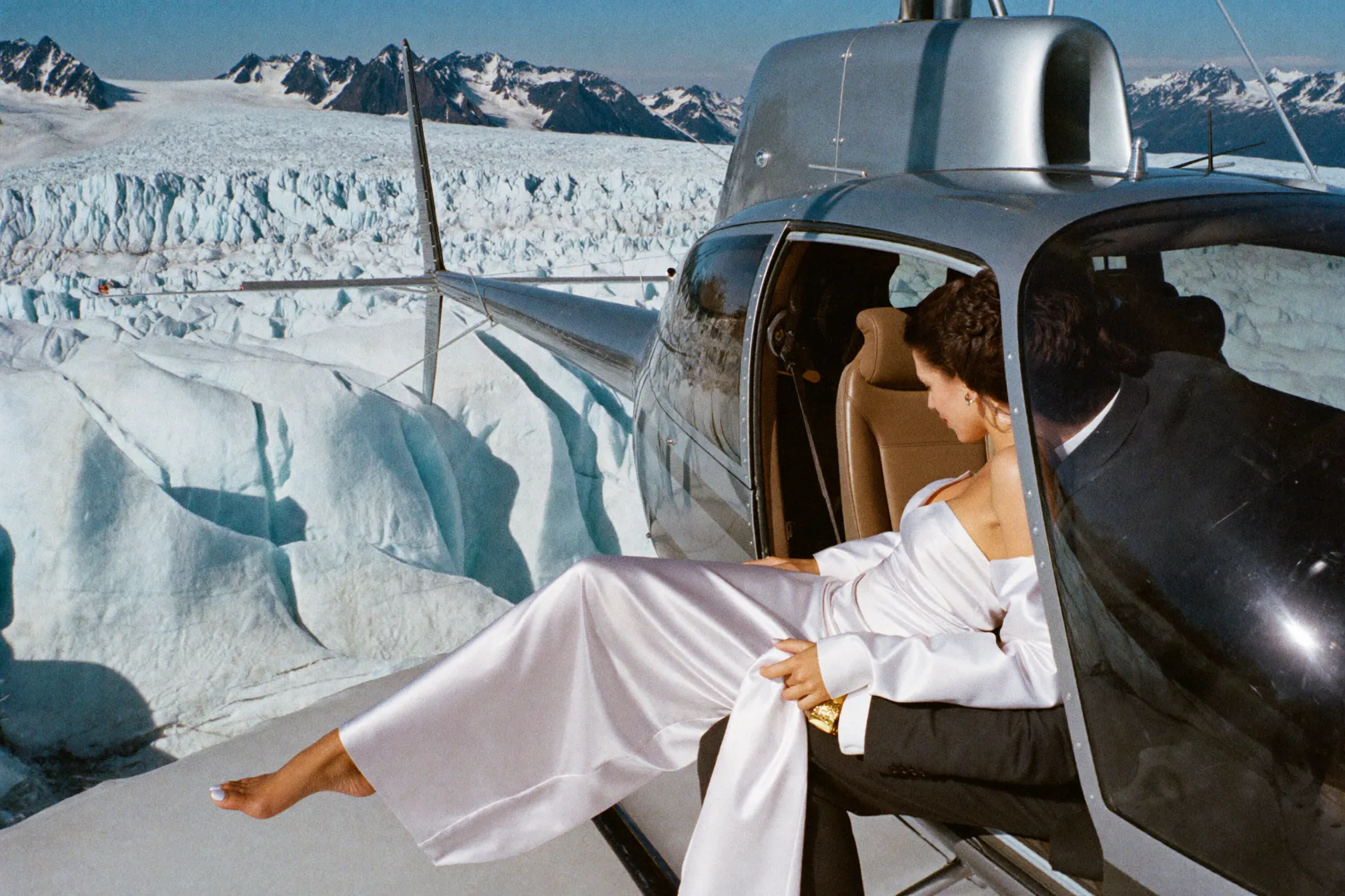 A couple stepping onto a remote snow-dusted alpine ridge beside a small helicopter